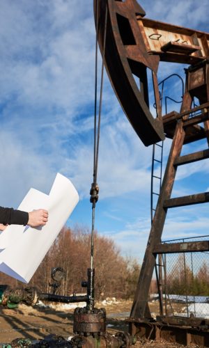 Side view of oil worker holding plan of oil field. Engineer in work uniform and helmet standing near petroleum pump jack under beautiful cloudy sky. Concept of petroleum industry and oil extraction.